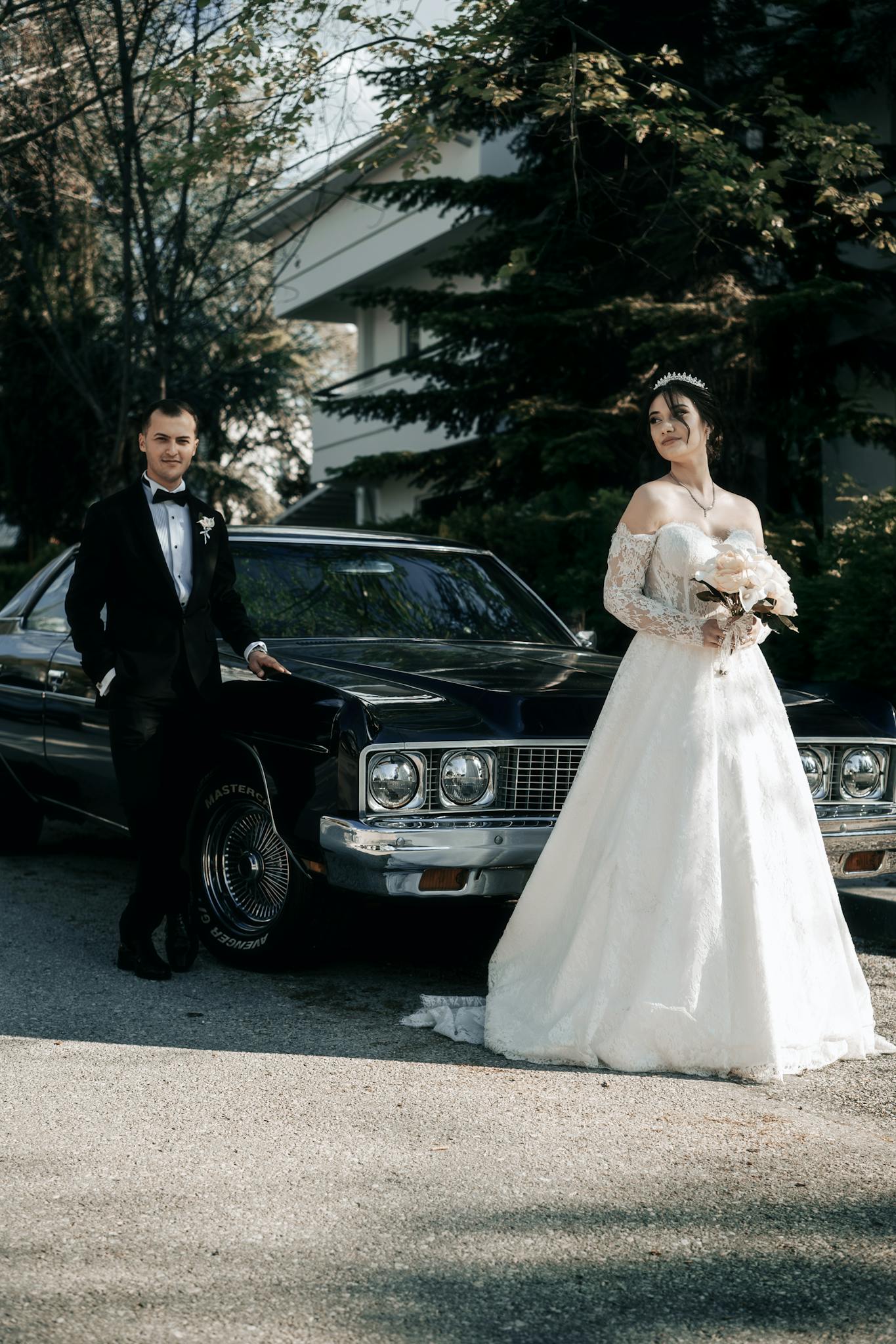 Bride and groom posing elegantly by a vintage car in an outdoor setting.